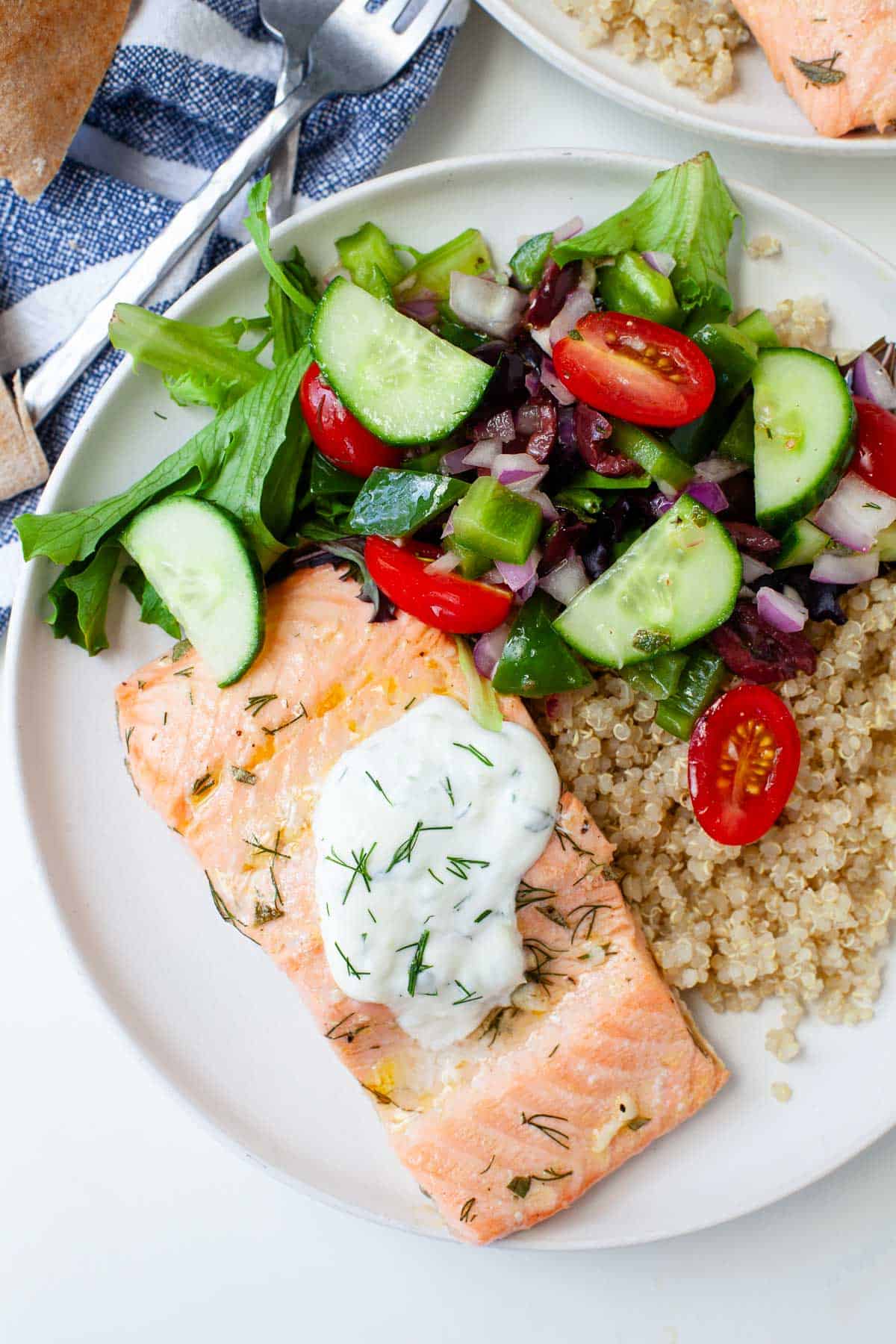 Greek salmon quinoa bowl on white dish with piece of salmon, tzatziki, quinoa, mixed greens, and chopped vegetables. 