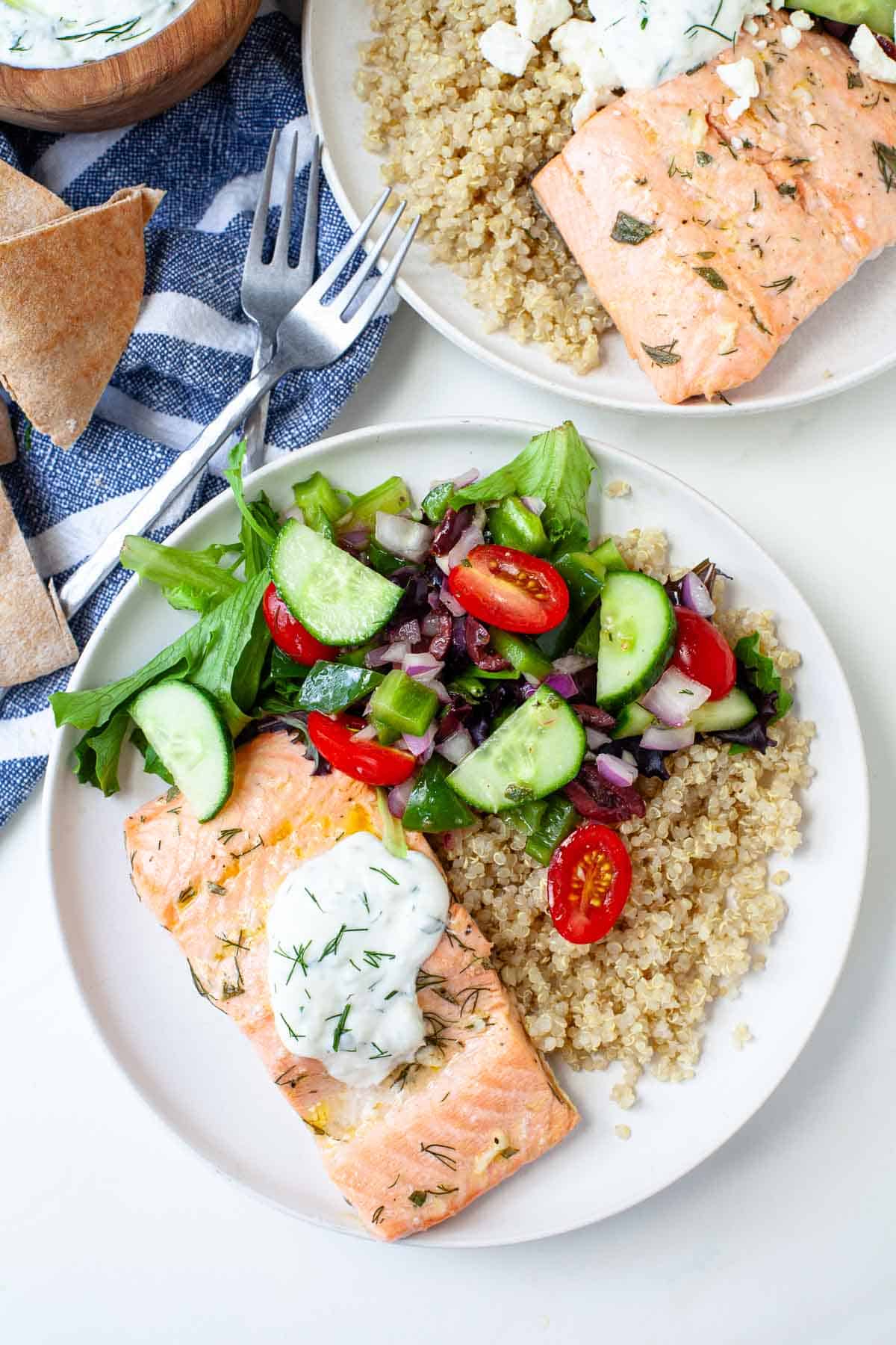 two Greek salmon quinoa bowls beside two silver forks, kitchen towel, pita bread and bowl of tzatziki. 