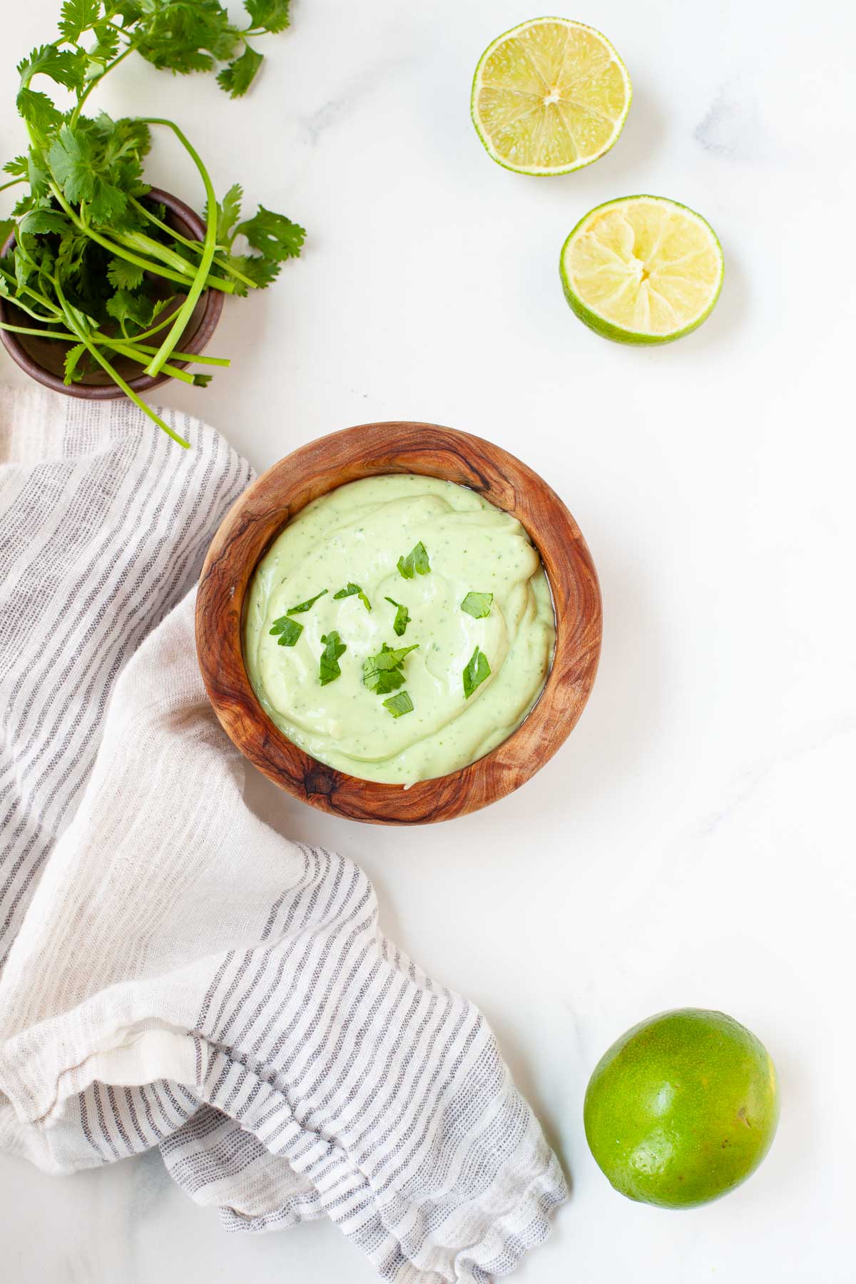 Avocado lime crema in wooden bowl beside kitchen towel, lime, and cilantro stems on white marble surface