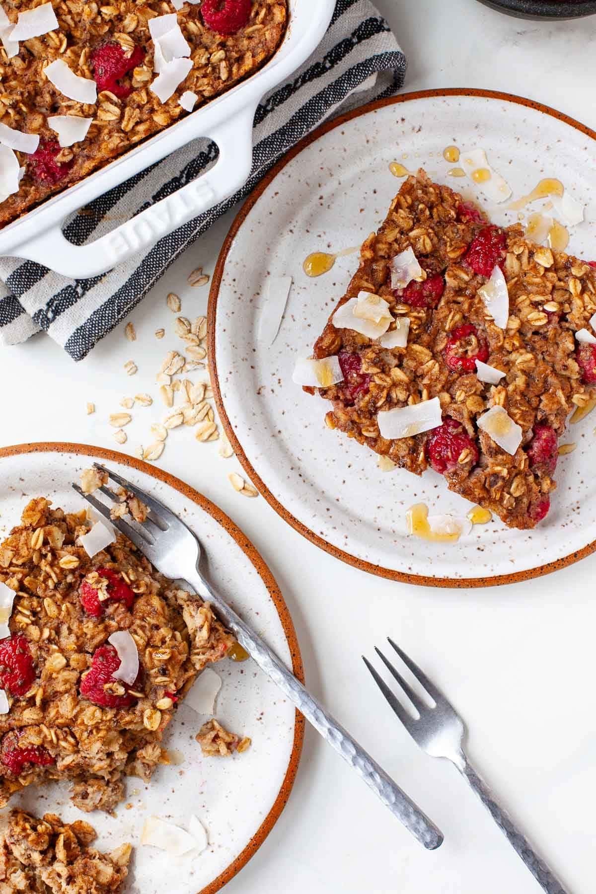 two plates with two pieces of baked oatmeal with raspberries, coconut flakes and syrup with two forks 