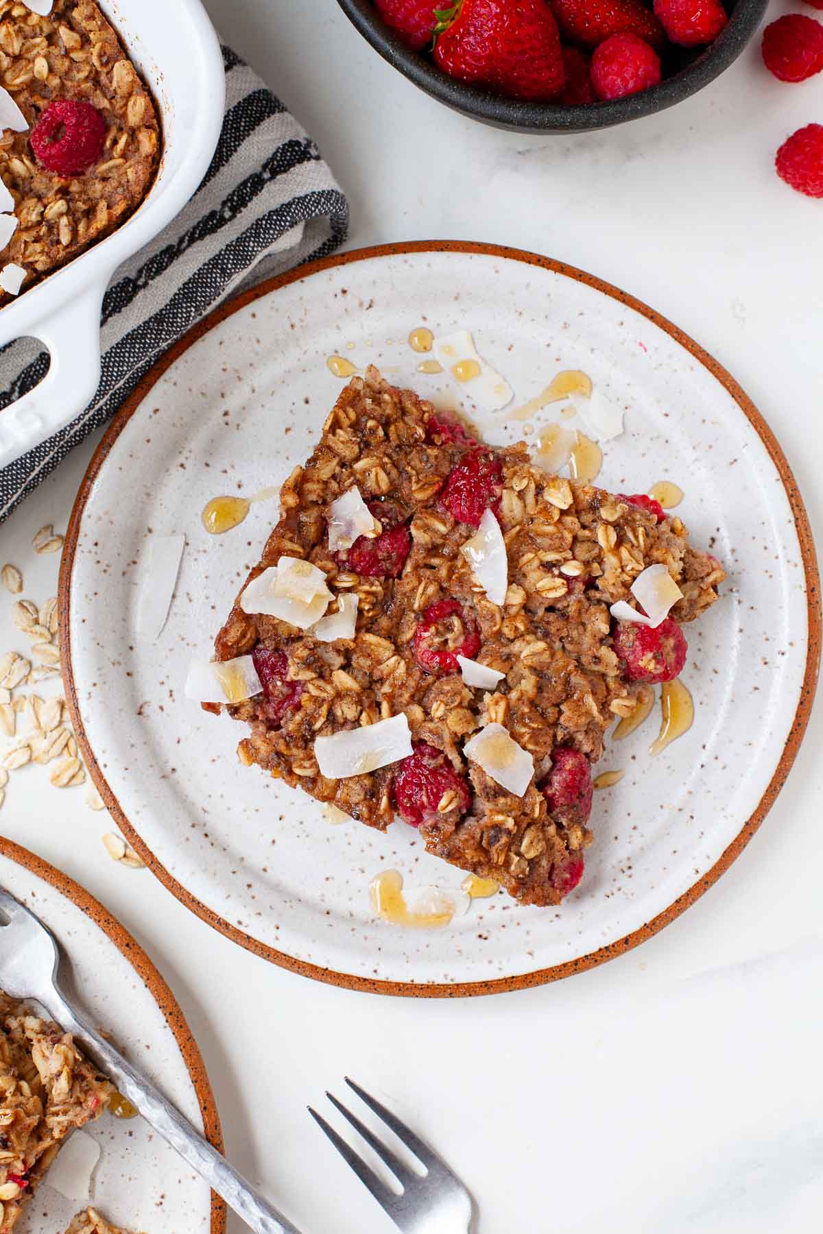 vegan baked oatmeal with raspberries and coconut flakes on white specked plate next to baking dish and bowl of berries