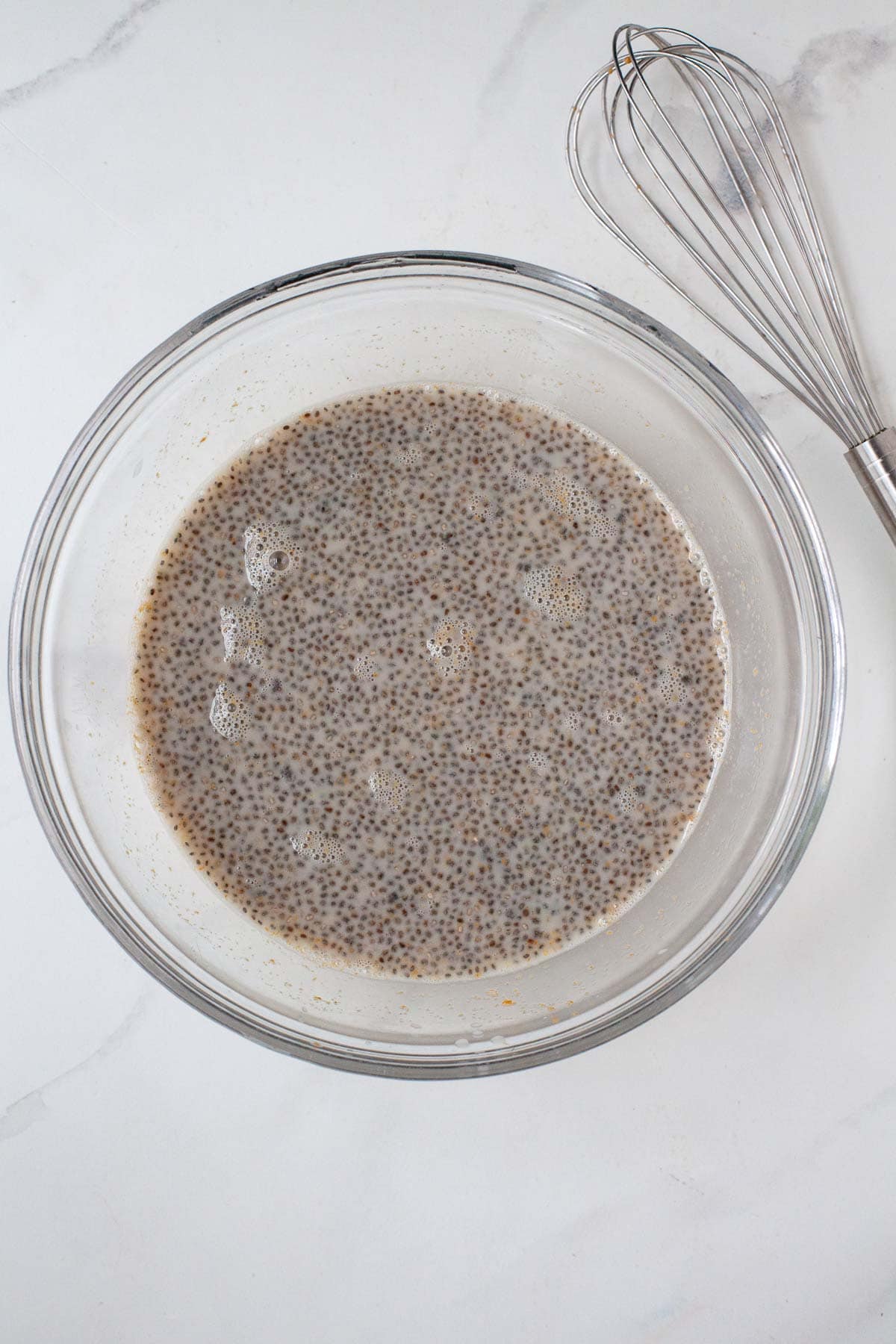 Combined ingredients wet ingredients for baked oatmeal including chia seeds in a large glass bowl next to whisk.