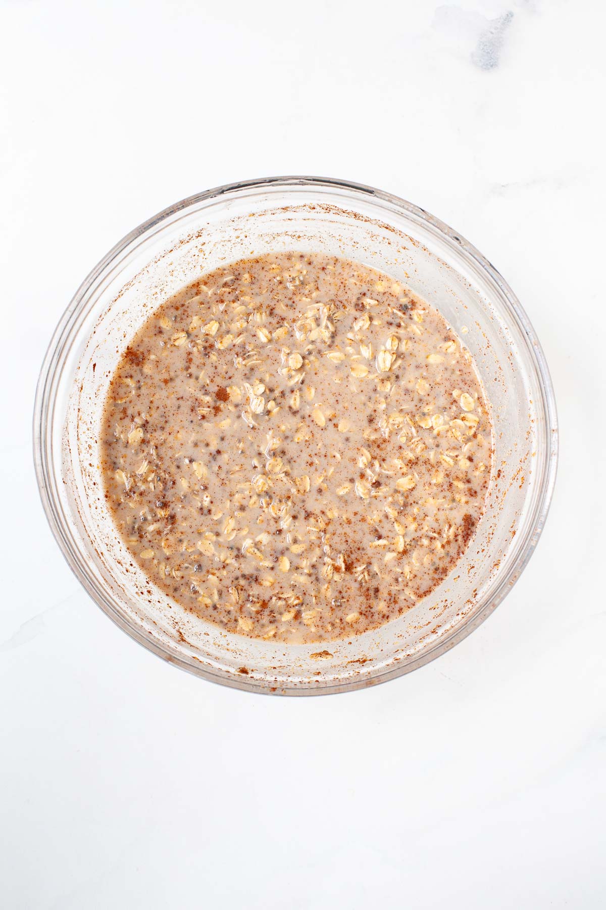 Rolled oats, baking powder, cinnamon, and salt mixed into wet ingredients in large glass bowl on marble counter.