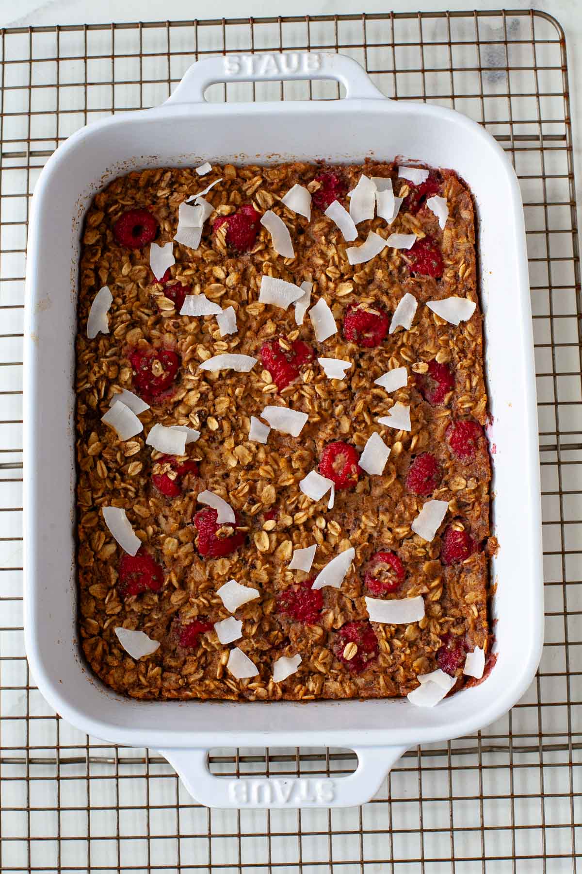 Baked vegan oatmeal topped with raspberries and coconut flakes in white ceramic baking dish on wire rack